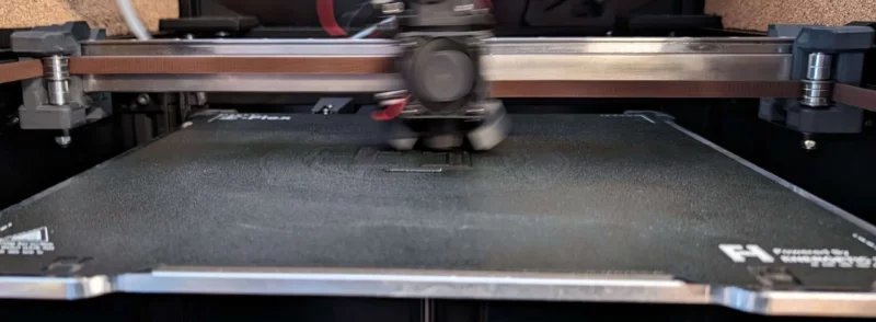 Toro-Titanium-Tube RatRig V-Core 4 3D printer printing a small part on a black build plate. Featuring a V-Core 4 with a moving print head.