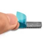 Close-up of a finger peeling protective blue film off a textured black nozzle brush for a 3D printer.